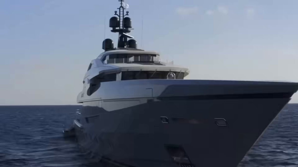 The image features a large yacht sailing through the ocean under a clear blue sky. The yacht has a white body with black accents, giving it an elegant appearance as it moves across the water. It's positioned towards the right side of the frame, creating a sense of motion and direction in the scene.