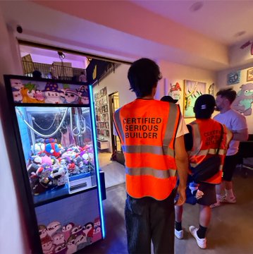 A photograph inside what appears to be a claw machine arcade showing a person wearing an orange 'CERTIFIED SERIOUS BUILDER' safety vest facing away from the camera, with a plush toy-filled claw machine on the left.
