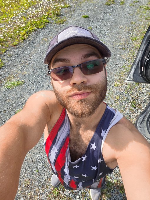 A selfie of a bearded man wearing a dark baseball cap, sunglasses, and an American flag tank top, taken outdoors near a gravel area with green grass.