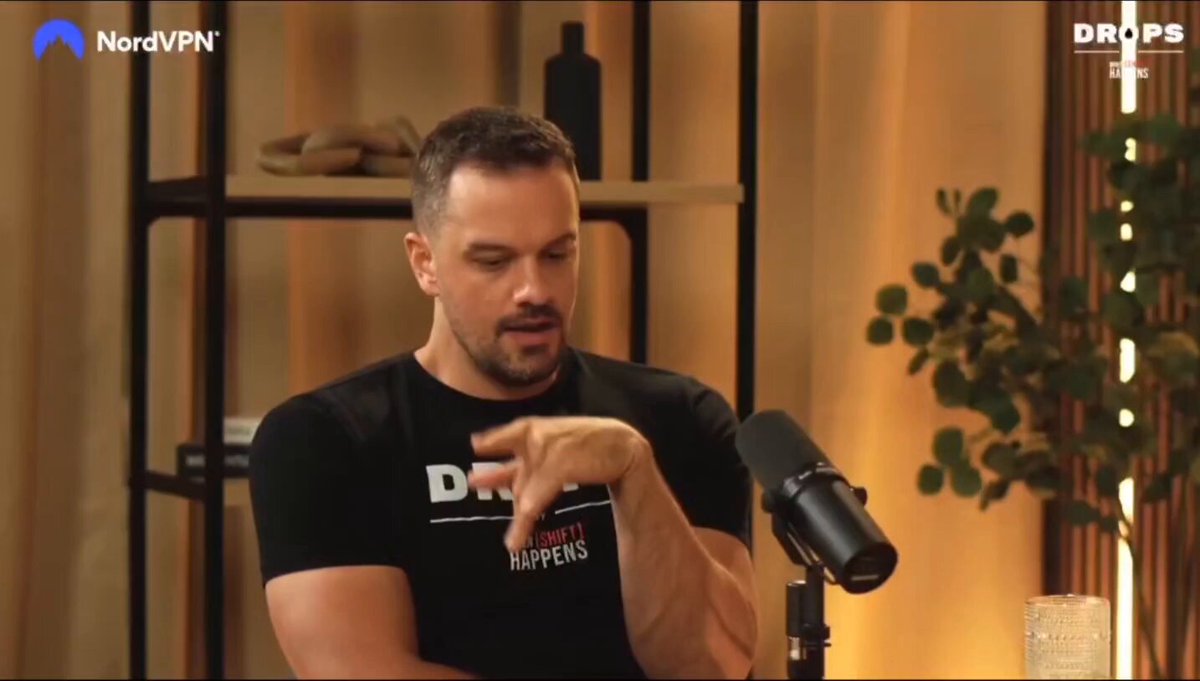 The image shows a man wearing a black t-shirt sitting at a desk with a microphone in front of him. The background features a bookshelf filled with various items, including several bottles and a potted plant. The caption for the image reads "Focus on what is in the image and how it relates to the post."