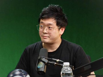 An Asian man with glasses speaking into a microphone in front of a green screen backdrop.