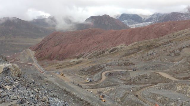 A winding dirt road and construction vehicles cut through the barren, red and grey slopes of a high-altitude mountain landscape, with peaks shrouded in clouds above an active mining site at 4,889m.