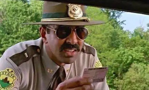 A state trooper wearing sunglasses and a hat examines a card, with a "STATE PATROL" patch visible on his shoulder, appearing to verify details relevant to the NFT "early alpha" claim in the accompanying text.