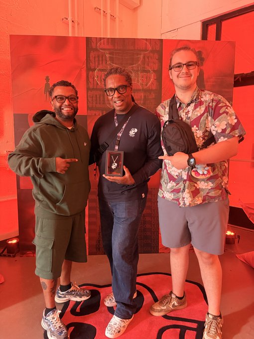 Three men stand together smiling at an event with red lighting; the man in the center holds a small award, and they are standing on a red carpet.