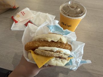 The image shows a person holding up a McDonald's breakfast sandwich, which includes a hamburger patty, cheese, and bacon. The sandwich is wrapped in paper and accompanied by a drink in a cup. The background of the photo features a dining table with various items on it, such as a red ketchup packet and a white napkin.