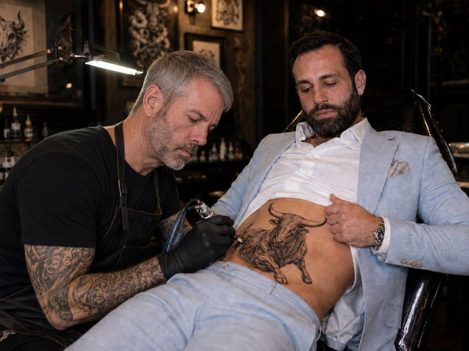 The image shows a tattoo artist with gray hair and tattooed arms working on a large bull tattoo on the torso of a seated client wearing a light blue suit, which visually represents the "Golden Bull Run" market rally referenced in the caption—suggesting that pulling out of the market now would be like getting a bullish tattoo only to abort midway.