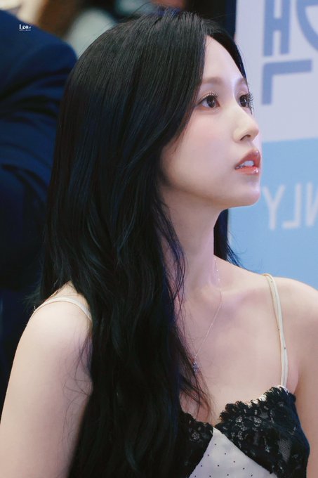 A young woman with long dark hair is shown in profile, wearing a black and white polka-dot top with lace trim and a delicate necklace, gazing to the side at what appears to be a media event. Behind her, partial white text on a blue backdrop reads "Y.J", consistent with the post identifying her as MINA from TWICE at a GMP event on July 21.