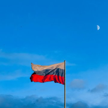 A Russian flag waves against a blue sky with the moon visible in the upper right corner, symbolizing the country referenced in the news about Russia's central bank publishing its first draft crypto market rules requiring digital depositories to hold up to $2.8M in capital ahead of a September 1 rollout.