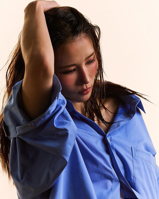 Portrait of a young woman with wet dark hair wearing an oversized light blue button-down Givenchy shirt, looking downward with her hand raised behind her head.
