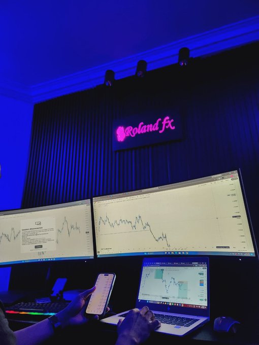 A dimly lit trading desk bathed in blue light with a neon pink "Roland fx" sign, featuring multi-monitor displays showing financial charts and a person holding a smartphone.