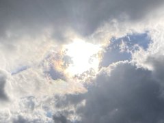 A photograph of a bright sun shining through gray clouds in a blue sky.