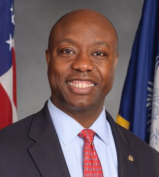 An official portrait of Senator Tim Scott, showing him smiling in a dark suit with a red tie, with American and other flags in the background.
