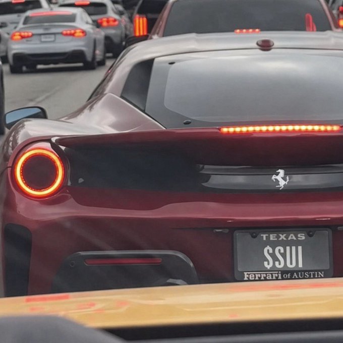 A red Ferrari sports car is shown from behind in traffic, featuring a Texas license plate that reads 'SSUI' with 'Ferrari of Austin' below it, alongside its iconic prancing horse logo.