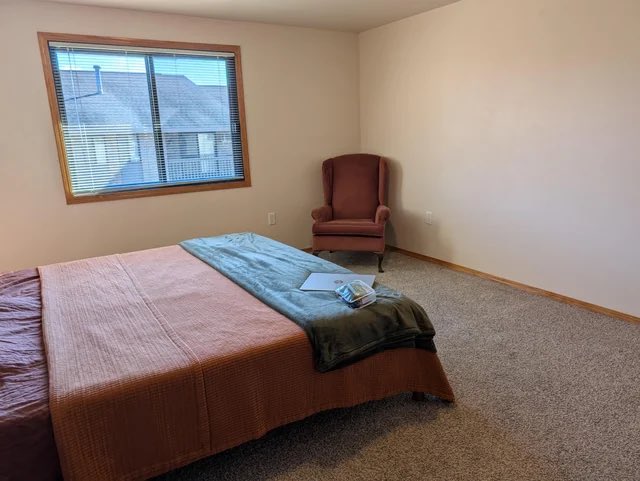 A sparsely furnished bedroom with a queen bed, pink and teal bedding, a pink wingback chair, and beige carpet sits below a window showing neighboring rooftops, with a small book or pamphlet resting on the bed. The image appears to be a listing photo, likely used to promote the room or home for sale or rent on the "pf app" (PostFinance app, as suggested by the Swiss-style architecture visible outside).