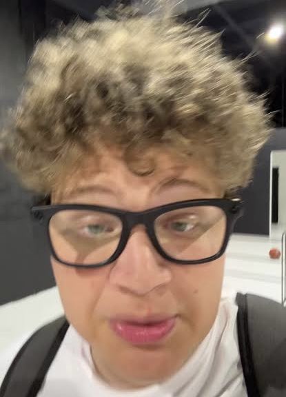 A close-up selfie of a person with light brown curly hair wearing thick black-framed glasses and pink lipstick.