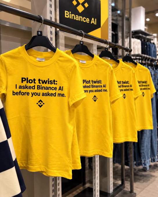 A row of yellow t-shirts hangs on a rack in a retail display, each printed with the text "Plot twist: I asked Binance AI before you asked me." beneath a Binance logo, with a "Binance AI" sign visible above.