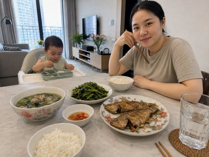 A woman and a baby in a high chair sit at a dining table featuring a home-cooked Vietnamese meal with fish, greens, soup, and rice.