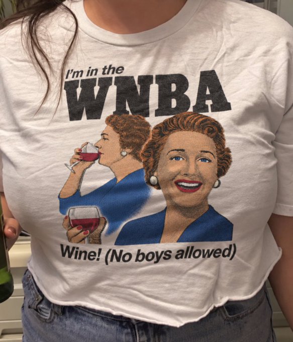 A person wearing a white t-shirt featuring a vintage-style illustration of two women drinking wine, with text reading 'I'm in the WNBA - Wine! (No boys allowed)'.