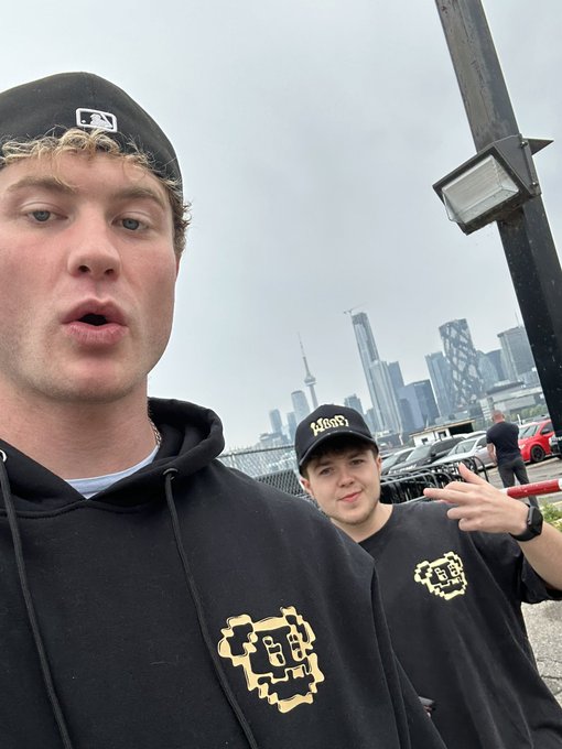 A selfie of two young men in black hoodies and caps, with the Toronto skyline, including the CN Tower, visible in the background.