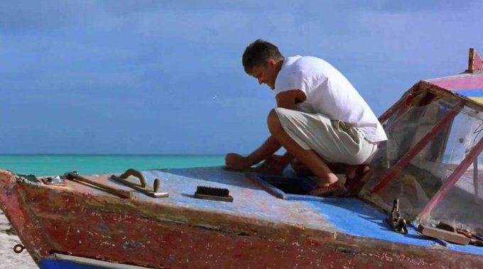 A man in a white shirt and beige shorts crouches barefoot on the deck of a weathered red and blue wooden boat, working with tools near the bow, with turquoise ocean water and a blue sky in the background.