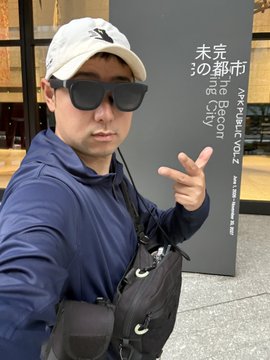 The image features a man wearing sunglasses, a hat, and a blue shirt with a black bag over his shoulder. He is posing for a photo near a building that has Japanese writing on it. The text "シンジくん、これが都市型仕様の第6型装備よ #STEPN" can be seen in the image, which might be related to an event or location with a Japanese theme.
