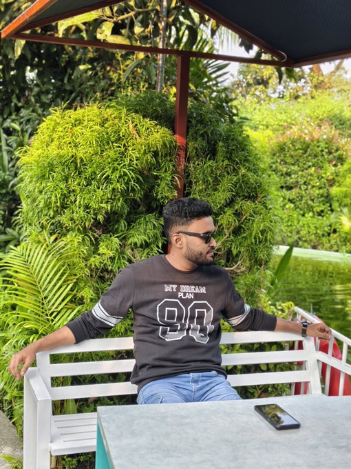 The image features a man sitting at an outdoor table under an umbrella, wearing a black sweatshirt with white text that says "90". He has his arm resting on the back of the bench while looking off into the distance. There are also two cell phones visible in the scene - one near the man and another further away from him. The background includes trees and bushes, creating a serene atmosphere for the outdoor setting.
