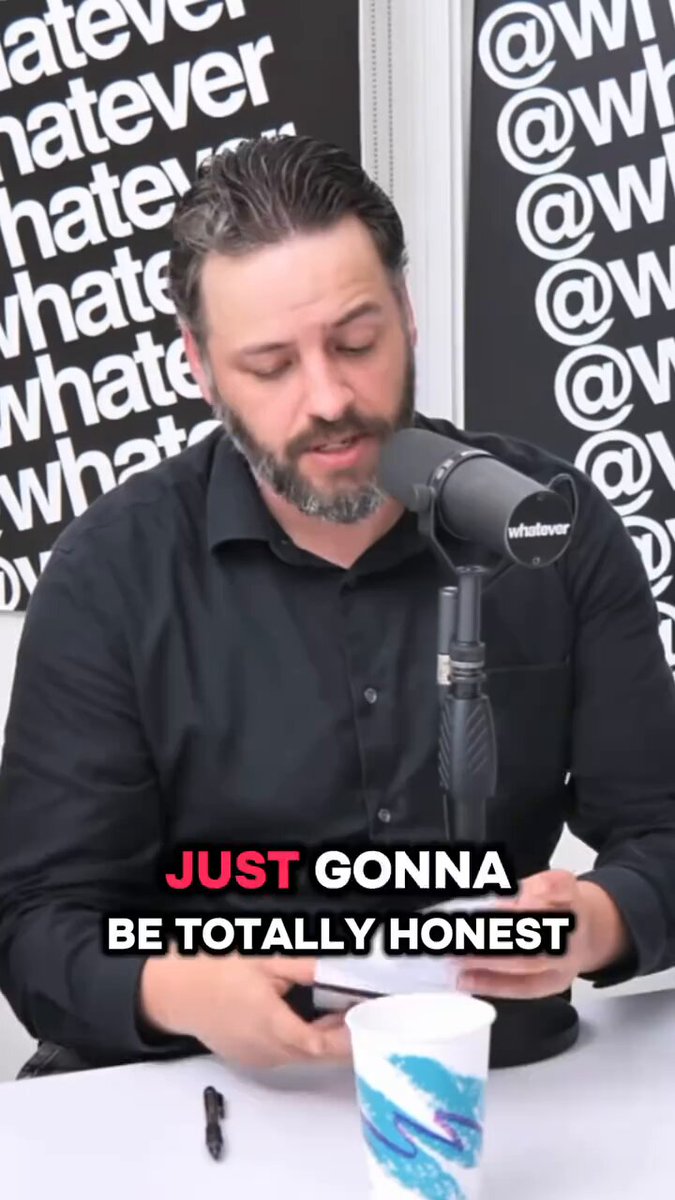 The image features a man sitting behind a microphone, wearing a black shirt with white text that reads "Just Gonna Be Totally Honest". The man appears to be talking about Bitcoin or cryptocurrency, as indicated by the presence of a cup on his desk. There is also a cup and a pen visible in the scene.