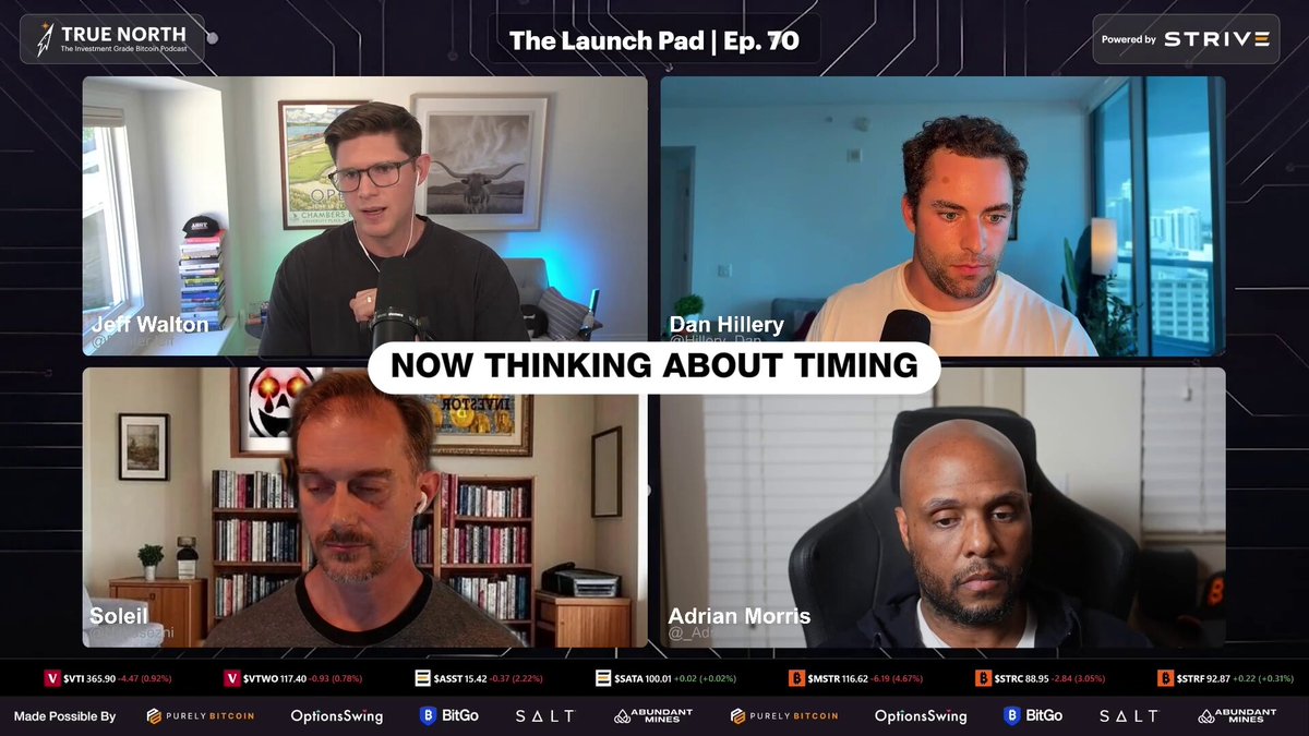 The image shows a group of four men sitting around a table, with one man speaking into a microphone while the others look at him attentively. The background features a computer screen displaying a video conference call between the four men. The caption for the image reads "Now thinking about timing."