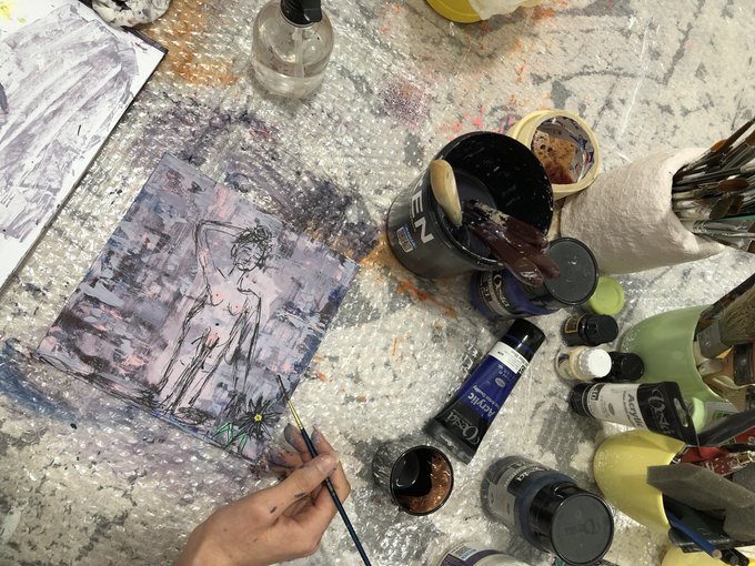 An overhead view of an artist's messy workspace, featuring a hand holding a thin brush over an ink sketch of a figure, surrounded by various paint tubes, brushes in holders, and jars of ink and pigment.