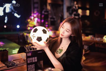 A young woman holding a soccer ball at what appears to be a sports bar or lounge setting, with Chinese text '6月开赛' visible on a sign.