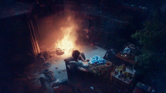 A dimly lit, messy room with a fire burning in the background and a person sitting on a couch in the foreground.