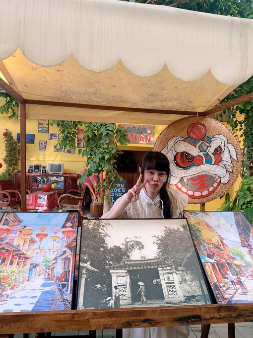 A young woman posing with a peace sign behind a display of vintage photographs at an outdoor café with a 'CỘC CỘC' sign, decorative plants, and a large traditional painted face decoration.