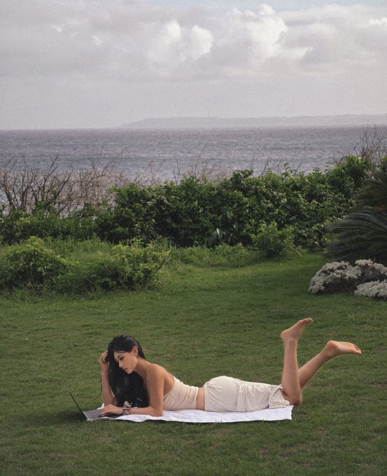 A woman with long dark hair lies on her stomach on a white blanket on a grassy hill overlooking the sea.