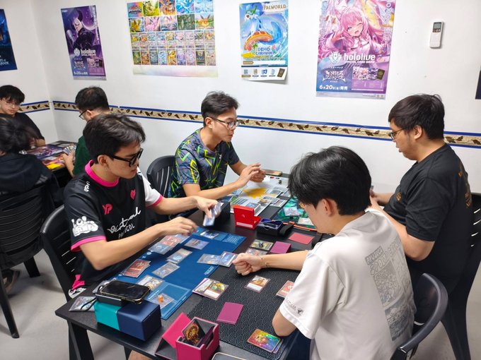 A group of young men sit around a table playing a trading card game in a room decorated with anime and gaming posters, including Hololive and Yu-Gi-Oh! banners.