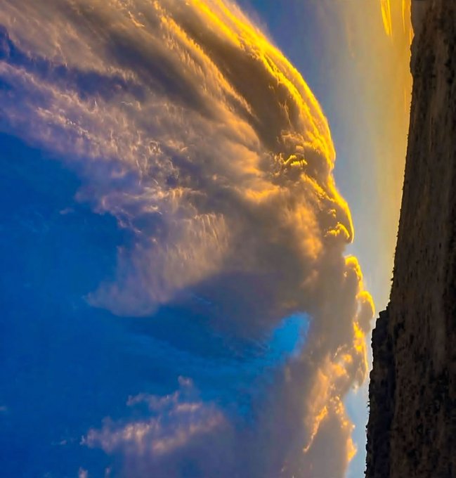 A sideways-oriented photograph captures a dramatic golden sunset illuminating fluffy clouds against a blue sky, with a dark textured edge on the right side.
