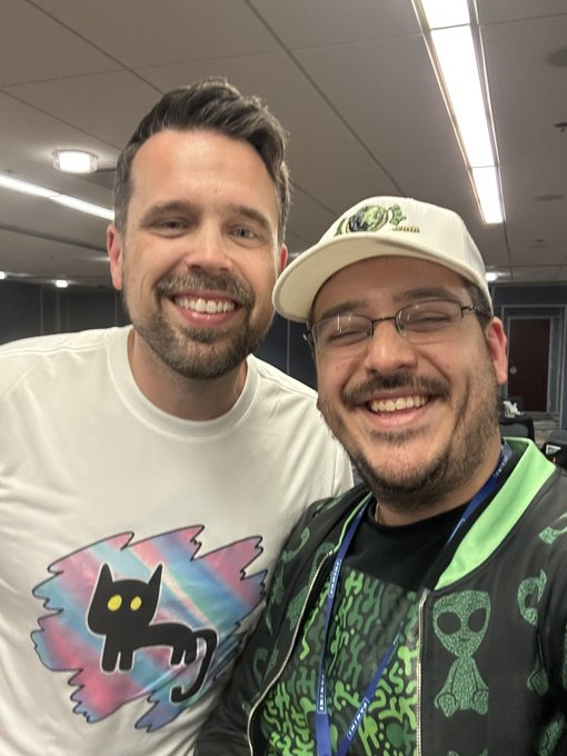 Two smiling men pose for a selfie in an office setting; one wears a white t-shirt featuring a black cat graphic with sunglasses, while the other wears a white cap and a green hoodie under a black jacket.