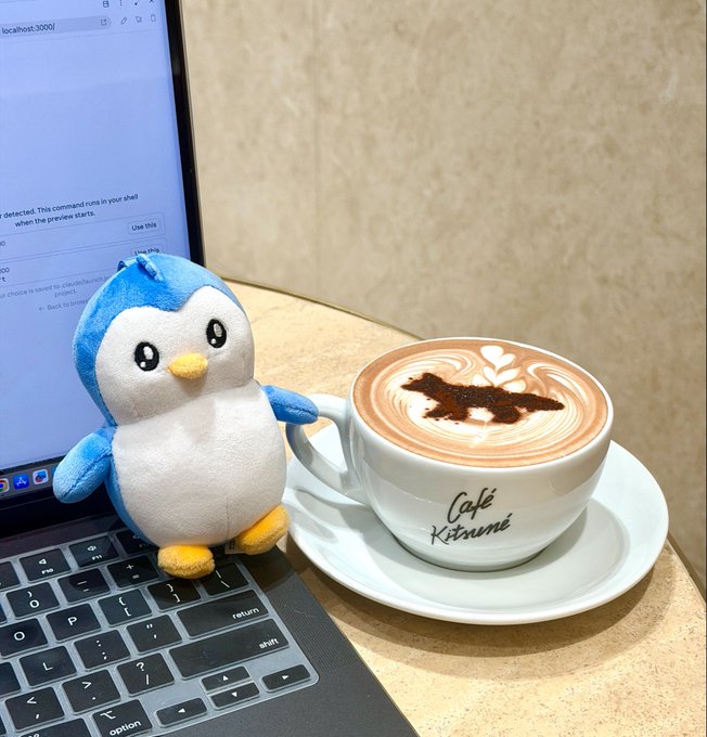 A plush penguin sits next to a Café Katsumi latte with leaf art on a saucer beside an open laptop displaying localhost:3000.