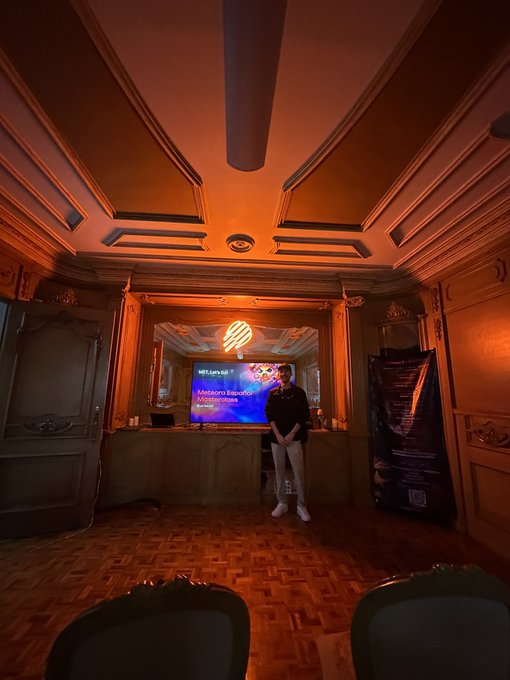 A young man stands on an ornately decorated stage with warm amber lighting and coffered ceilings, appearing honored while speaking at an event. A screen beside him displays the text "Not a Spot Token" along with what appears to be LP-related messaging, reflecting his role as a community host and LP Army member.
