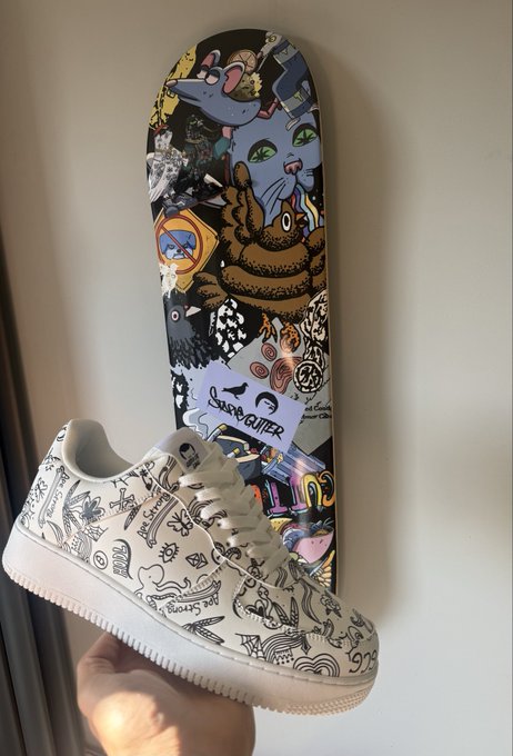 A hand holding a white sneaker with black doodle designs, positioned in front of a skateboard deck covered in colorful stickers and graphics.