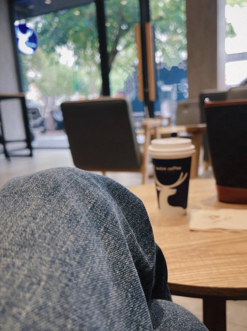 A first-person view of someone in a coffee shop, looking past their denim-clad knee at a wooden table with a Luckin Coffee cup, with a street scene visible through the window.