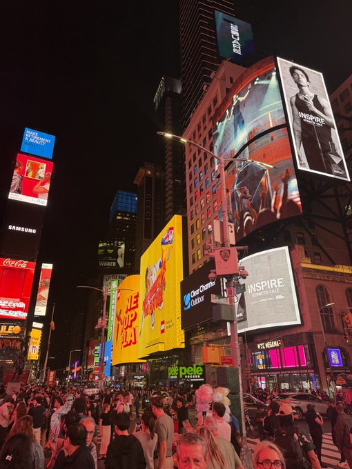 A bustling Times Square in New York City at night, crowded with pedestrians and lit up with bright digital billboards featuring advertisements for Coca-Cola, Samsung, McDonald's, and 'INSPIRE' shows.