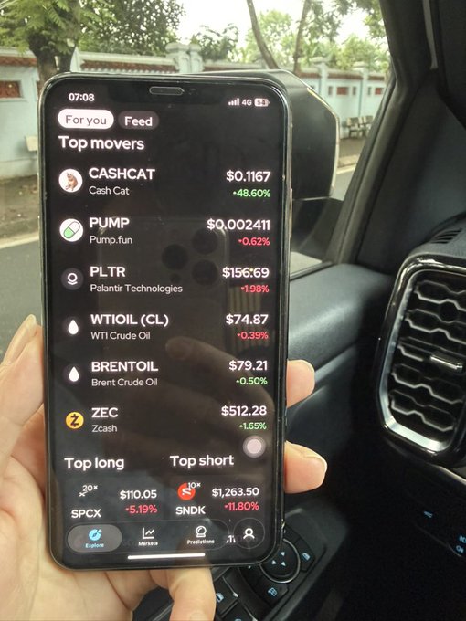A hand holds a smartphone inside a car displaying a stock/crypto trading app with 'Top movers' including CASHCAT (+48.60% at $0.1167), PUMP (-0.62%), PLTR (+1.98% at $156.69), WTIOIL (CL), BRENTOIL, and ZEC (+1.65%).