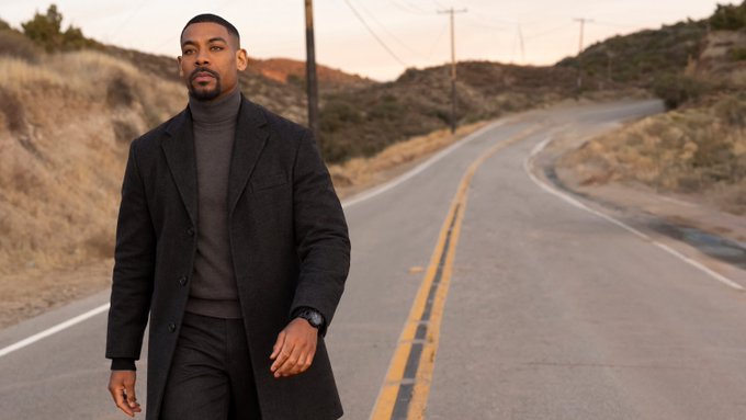A well-dressed man wearing a dark coat and gray turtleneck walks along an empty desert highway with power lines stretching into the distance.