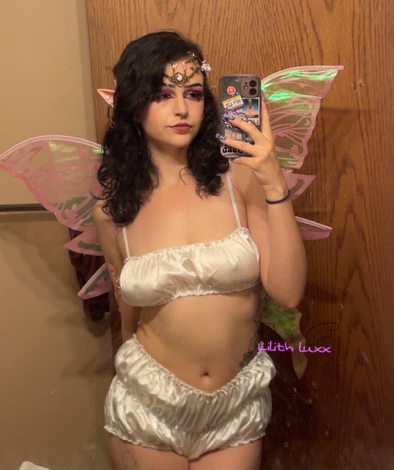 A person with dark curly hair, wearing pink and green fairy wings, a jeweled headpiece, and white satin lingerie, takes a mirror selfie with a phone case full of stickers; text 'Lilith luxx' appears in pink.