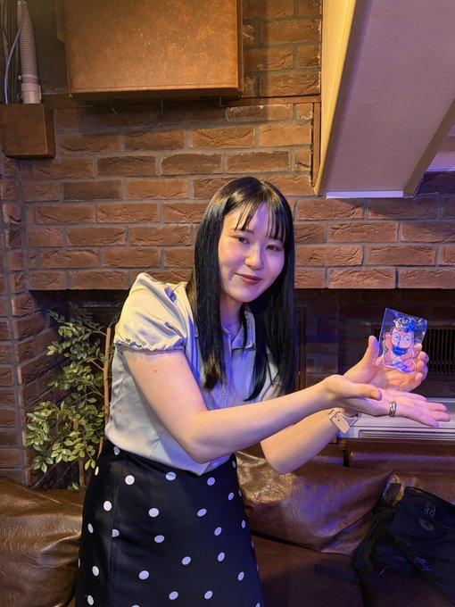 A young woman with long black hair, wearing a light blouse and black polka-dot skirt, smiles while displaying a Pokémon trading card (Gengar) in a warmly lit room with brick walls.