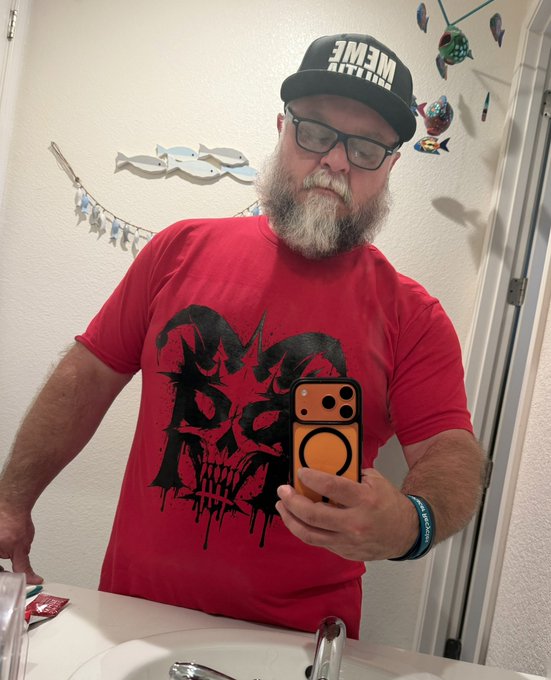 A bearded man wearing glasses, a backwards 'MEME' cap, and a red shirt with a black skull graphic takes a mirror selfie with an orange phone in a bathroom.