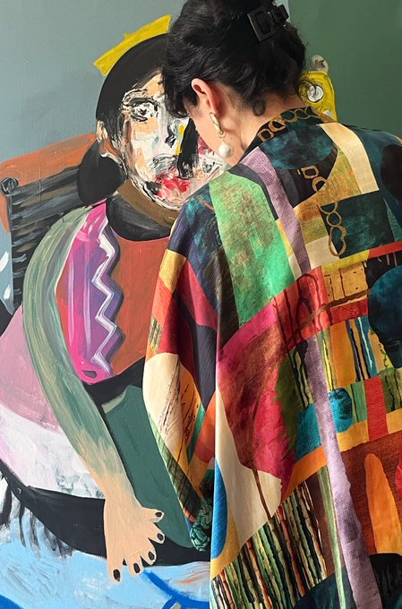 A woman with dark hair in a ponytail stands with her back to the camera, wearing a vibrant, multicolored patterned coat, while gazing at a large abstract portrait painting on the wall. The bold, expressive artwork echoes the artistic and eclectic energy of her colorful attire, setting a creative and inspiring tone for the "Good Morning Everyone" greeting.