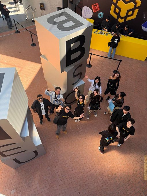 A group of people wave enthusiastically from below giant cube-shaped displays featuring the letters "BNB" at what appears to be the #BNB Seoul event, with staff in black "BNB" shirts helping manage the crowd. The scene captures the lively atmosphere described in the post, showing event operations and the packed, successful gathering of BNB Chain ambassadors and supporters.