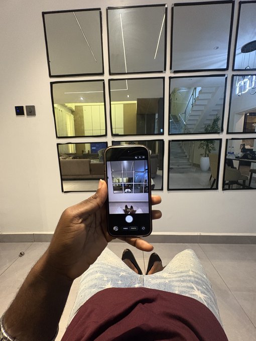 A person takes a first-person selfie with their phone, capturing their own reflection in a grid of mirrored wall panels while sitting down. The image's focus on self-reflection visually complements the post's casual "Gm how are we making money today?" greeting, suggesting a moment of self-assessment before tackling financial goals.