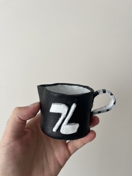 A hand holds up a small black ceramic mug with "ZL" painted in white lettering. The mug appears to be a handcrafted, artisan-style piece, likely representing the "$ZL" ticker token referenced in the post about supporting and showcasing overlooked projects in the crypto community.